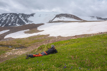 Man photographing orange trollius flowers on green grass with view to large snowy mountain dome under cloudy sky. Tourist with camera against high snow mountain in dome shape at changeable weather.の写真素材