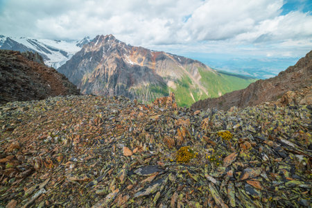 Vivid mountain landscape near precipice edge with sharp stones with view to sunlit large pinnacle with snow under cloudy sky. Picturesque view from stone hill to mountain peak with shadows from cloudsの写真素材