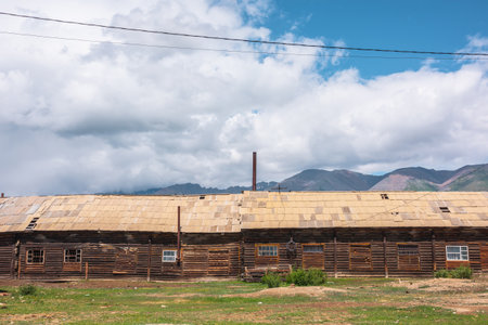 Large long wooden barn with sloping roof in sunlight under cloudy sky at changeable weather in mountains. Sunlit old wide wood house under wires against high rocky sharp mountain range in low clouds.の写真素材