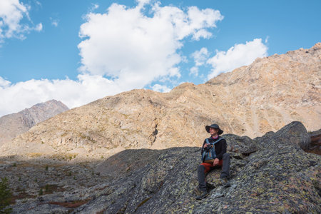 Hiker with backpack sitting on stone against rocks in bright sunlight. Alone man with photo camera in bright sun near sunlit rocky mountain wall. Tourist on moraines in high mountains in sunny day.の写真素材