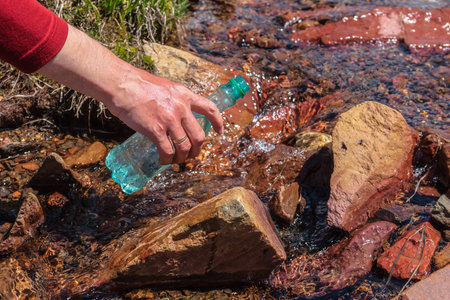 Man replenishes water from mountain stream in bright sun. Tourist fills bottle with mineral water in sunny day. Hand with bottle in clear water stream in sunlight. Replenishment of water during hike.の写真素材