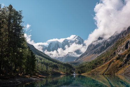 Scenic landscape with turquoise mountain lake in autumn valley against large snow mountains in low clouds in morning sunlight. Alpine lake with view to sunlit high snowy mountain range in low clouds.の写真素材