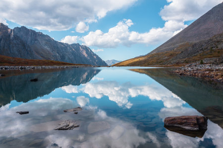 Tranquil autumn landscape with clouds reflection on smooth mirror surface of mountain lake in high hanging valley. Meditative view from calm alpine lake to mountain vastness. Stones in clear water.の写真素材