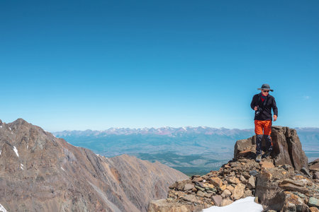 Tourist walks on mountain near abyss edge on high altitude under blue sky in sunny day. Man with camera on high rock near precipice edge with wonderful view from above to large mountain range in away.の写真素材