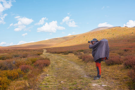 Alone traveler in raincoat with large backpack photographs nature among fading autumn vegetations on high mountain plateau. Backpacker with photo camera shoots mountain landscape in changeable weatherの写真素材