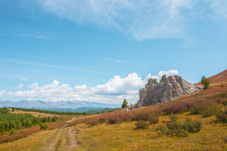 Atmospheric autumn landscape with big rock on hill with view to sunlit mountain vastness and high mountain range line on horizon under clouds. Impressive mountain scenery in vivid fading autumn colorsの写真素材