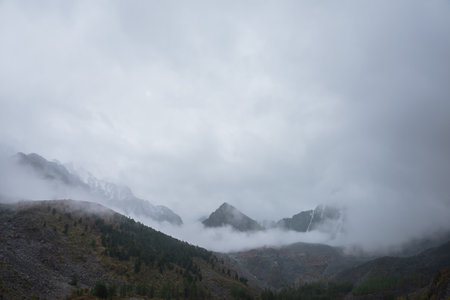 Dark atmospheric landscape with high mountain silhouettes in dense fog in rainy weather. Snowy rocky mountain top above hills in thick fog in dramatic overcast. Black rocks in low clouds during rain.の写真素材