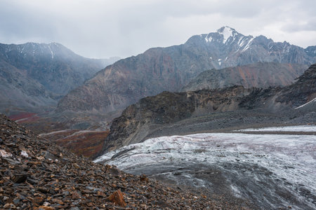 Awesome landscape with long glacier tongue against high mountain range with sharp rocky peaked top in overcast weather. Cone shaped pointy peak in dramatic cloudy sky. Large glacier among sharp rocks.の写真素材