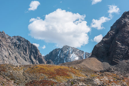 Big cloud in blue sky above large snow mountain top with glacier in autumn sunny day. Vivid autumn colors in high mountains. Motley landscape with sharp rocks and snowy mountain peak in bright sun.の写真素材