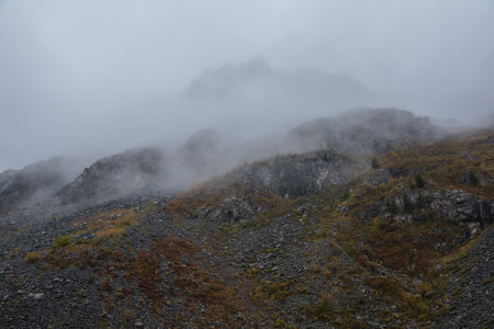 Dark atmospheric landscape in fading autumn colors with high rocky hill and silhouette of sharp rocks on mountain top in dense fog in rainy weather. Large black peak in thick low clouds during rain.の写真素材