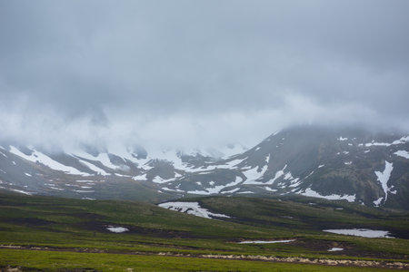Dramatic alpine landscape with snowy mountains in gray low clouds. Bleak atmospheric scenery of tundra under lead gray sky. Gloomy minimalist view to mountain range among low rainy clouds in overcast.の写真素材
