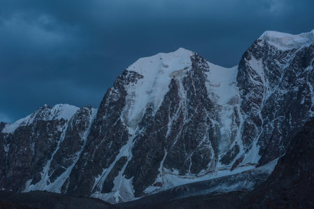 Atmospheric nightly landscape with huge snowy mountain top in dramatic sky. Hanging glacier and cornice on beautiful giant snow mountains in night. High snow-covered mountain range in dusk dim light.の写真素材