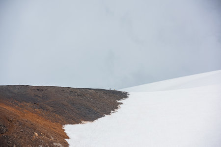 Scenic alpine landscape with small people on high snowy mountain in sunlight under gray cloudy sky. Awesome mountain scenery with small tourists on sunlit large snow mountain at changeable weather.の写真素材