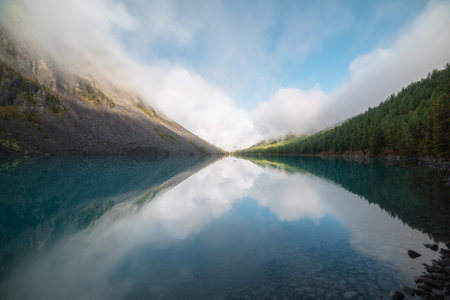 Scenic view to big pure mountain lake in forest valley and low clouds above firs in horizon. Lovely tranquil meditative landscape with low clouds reflection in turquoise glacial lake in early morning.の写真素材