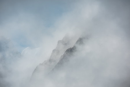 Lovely scenery with big snowy mountains in thick clouds. High mountain wall in gantly cloudy sky. Scenic view to large snow mountain in clearance of dense fog. Beautiful diagonal rocks in low clouds.の写真素材