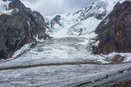 Dramatic landscape with glacier icefall on large snow mountain range with sharp rocky pinnacle under gray cloudy sky. Big glacier close-up in high altitude. Gloomy scenery in mountains in overcast.の写真素材