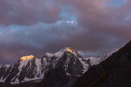 Atmospheric landscape with sunset gold reflection on huge snowy mountain top in violet dramatic sky. Scenic view to giant snow mountains in dusk. Snow-covered mountain range silhouettes in twilight.の写真素材