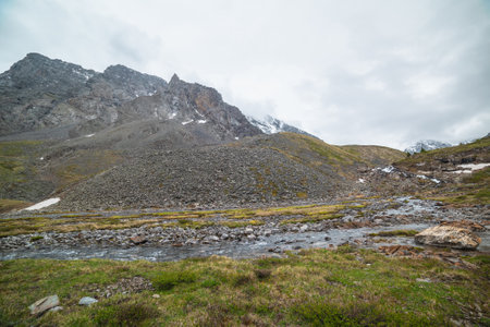 Atmospheric landscape with clear mountain stream from hills against snowy mountain range with peaked top in low clouds. Beautiful mountain creek and high sharp rocks under cloudy sky in gloomy weatherの写真素材