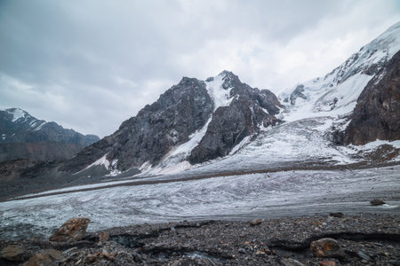 Dramatic landscape with glacier tongue and icefall on large snow mountain range with sharp rocky pinnacle under gray cloudy sky. Long glacier in high altitude. Gloomy scenery in mountains in overcast.の写真素材