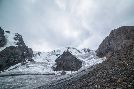 Dramatic landscape with two glacier icefalls on large snow mountain range with sharp rocks under gray cloudy sky. Long glacier with icefall in high altitude. Gloomy scenery in mountains in overcast.の写真素材