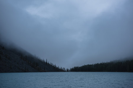 Dark atmospheric landscape with ripples on big blue mountain lake and black pointy firs silhouettes in low clouds. Tranquil view to deep blue alpine lake and peaked tops of spruces in low cloudy sky.の写真素材
