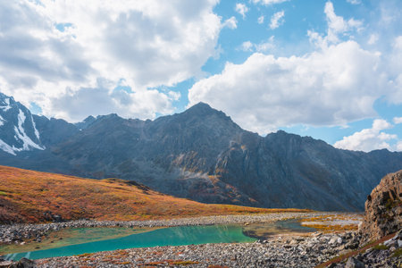 Lovely autumn landscape with turquoise mountain lake against high mountain top in sunlight under clouds in blue sky. Sunlit beautiful small alpine lake and large mountain range. Vivid autumn colors.の写真素材