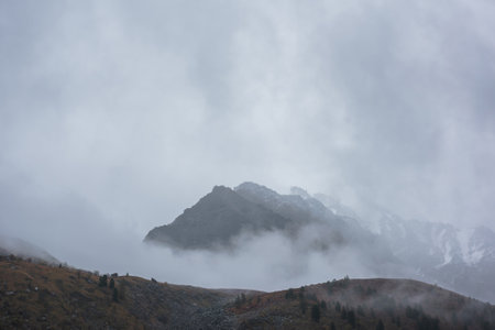 Dark atmospheric landscape with high mountain silhouettes in dense fog in rainy weather. Snowy rocky...