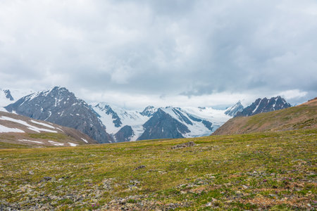 Scenic alpine view from sunlit green grassy hill to high snowy mountain range with sharp tops and glaciers under gray cloudy sky. Colorful landscape with large snow mountains at changeable weather.の写真素材