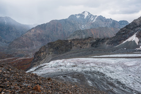 Awesome landscape with long glacier tongue against high mountain range with sharp rocky peaked top in overcast weather. Cone shaped pointy peak in dramatic cloudy sky. Large glacier among sharp rocks.の写真素材