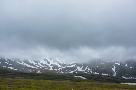 Dramatic alpine landscape with snowy mountains in gray low clouds. Bleak atmospheric scenery of tundra under lead gray sky. Gloomy minimalist view to mountain range among low rainy clouds in overcast.の写真素材