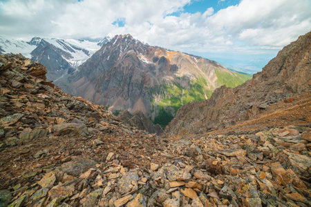 Vivid mountain landscape near precipice edge with sharp stones with view to sunlit large pinnacle with snow under cloudy sky. Picturesque view from stone hill to mountain peak with shadows from cloudsの写真素材
