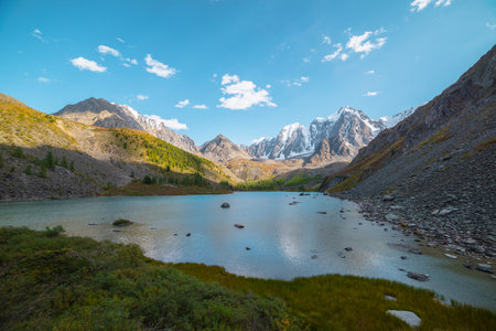 Pure azure alpine lake with view to forest hill and giant snow mountains in autumn sunny day....