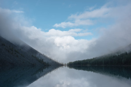 Tranquil meditative misty scenery of glacial lake with reflection of pointy fir tops and clouds at early morning. Graphic EQ of spruce silhouettes on calm alpine lake horizon. Mountain lake in fog.の写真素材