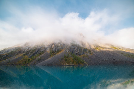 Pure mountain lake against sunlit sharp rocks in low clouds. Tranquil meditative landscape with...