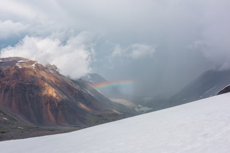 Alpine landscape with vivid rainbow above mountain lake. Scenic view from snow hill to bright rainbow above lake in mountain valley. Top view from snow mountain to colorful rainbow and low clouds.の写真素材