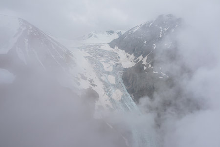Atmospheric mountain landscape with long vertical glacier tongue with cracks among rocks in dense low clouds. Awesome aerial view to large glacier with icefall in thick low clouds in high mountains.の写真素材