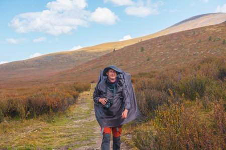 Alone traveler in raincoat with large backpack walks along hiking trail among fading autumn vegetations on high mountain plateau. Backpacker with photo camera in mountain trekking in rainy weather.の写真素材