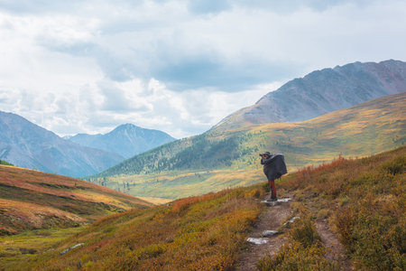 Alone traveler in raincoat with large backpack photographs nature among fading autumn vegetations on hiking trail through mountain pass. Backpacker with photo camera shoots autumn mountain landscape.の写真素材