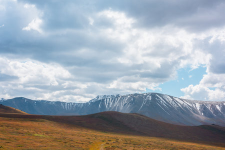 Motley autumn landscape with hills on high plateau and sunlit snowy mountain range under dramatic cloudy sky. Vivid autumn colors in mountains. Sunlight and shadows of clouds in changeable weather.の写真素材