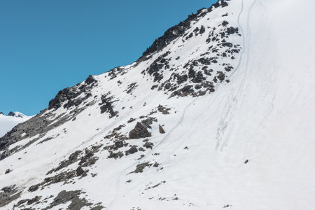 Climbers footprints on steep snowy slope of high snow mountain with rocks on top. Snow-white mountain under blue sky. Minimal landscape with snowy mountain with alpinists footprints at high altitude.の写真素材