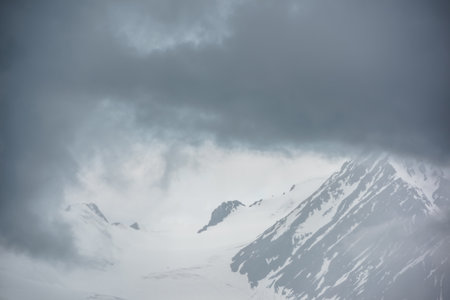 Atmospheric mountain landscape with fuzzy silhouettes of rocks on glacier in low clouds during rain. Dramatic view to large glacier tongue in snow mountains blurred in rain haze in gray low clouds.の写真素材
