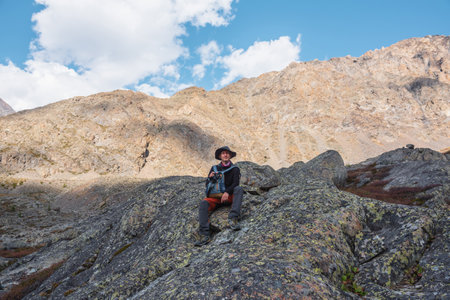 Hiker with backpack sitting on stone against rocks in bright sunlight. Alone man with photo camera in bright sun near sunlit rocky mountain wall. Tourist on moraines in high mountains in sunny day.の写真素材