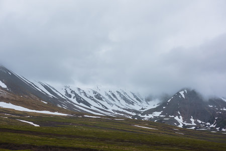 Dramatic alpine landscape with snowy mountains in gray low clouds. Bleak atmospheric scenery of tundra under lead gray sky. Gloomy minimalist view to mountain range among low rainy clouds in overcast.の写真素材