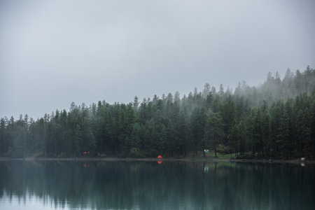 Tranquil atmospheric scenery with tents near mountain lake among coniferous trees silhouettes in dense fog. Pure alpine lake in mystery thick fog. Glacial lake and forest edge in misty early morning.の写真素材