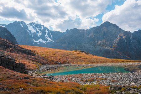 Lovely autumn landscape with turquoise mountain lake against high snowy mountain range in sunlight under clouds in blue sky. Sunlit beautiful small lake and large snow mountains. Vivid autumn colors.の写真素材