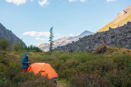 Tourist with photo camera near orange tent among lush vegetation in valley in sunny day. Man resting in nature during autumn mountain hike in good weather. Photographer takes pictures of wild nature.の写真素材