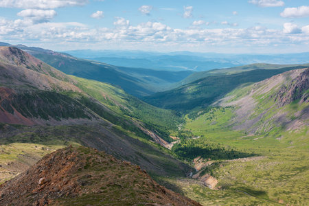 Scenic aerial view from mountain pass to green forest valley among mountain ranges and hills on horizon at changeable weather. Green landscape with sunlit mountains under cumulus clouds in blue sky.の写真素材