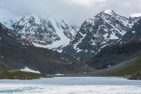 Dramatic mountain landscape with frozen alpine lake and high snow mountains and sunlit big rocks among low clouds. Atmospheric scenery with ice lake on background of large snowy mountains in overcast.の写真素材