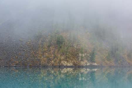 Simple meditative landscape with forest hill reflection in tranquil turquoise lake in fog. Mossy shore with firs in fading autumn colors reflected in mirror mountain lake. Autumn shore of alpine lake.の写真素材
