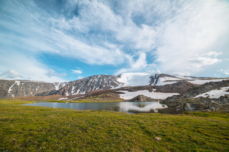 Colorful green landscape with mountain lake in sunlit meadow against high snowy mountain range under cloudy sky in changeable weather. Vivid sunny scenery with snow mountains reflected in alpine lake.の写真素材